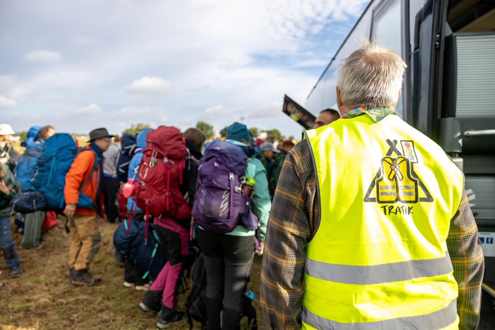 Frivillig i gul refleksvest med trafik-tryk tager imod spejdere med rygsække ved bussen på Spejdernes Lejr 2026